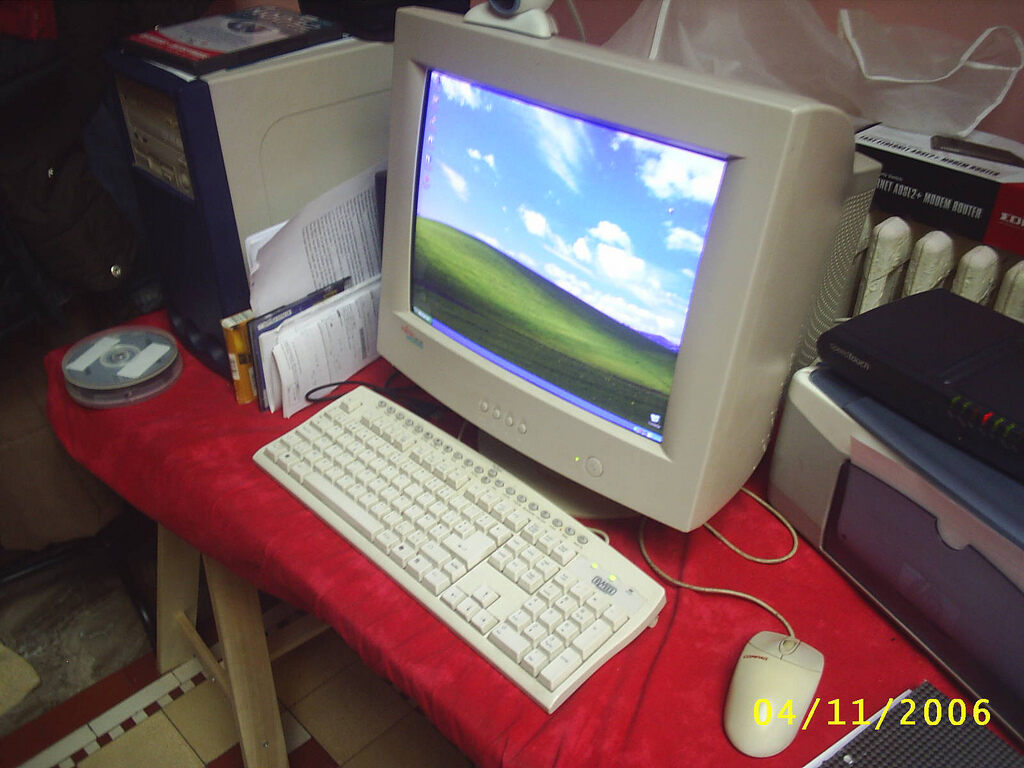 Um computador com monitor CRT exibindo o papel de parede padrão do Windows XP: uma colina verdejante sob o céu azul